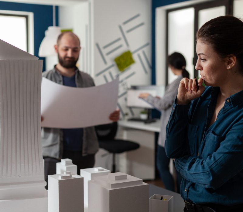 Architect in modern office thinking about design work standing next to tall building foam model Architect in modern office thinking about design work standing next to tall building foam model with team colleague engineer holding blueprints. Team of architects doing creative thinking.